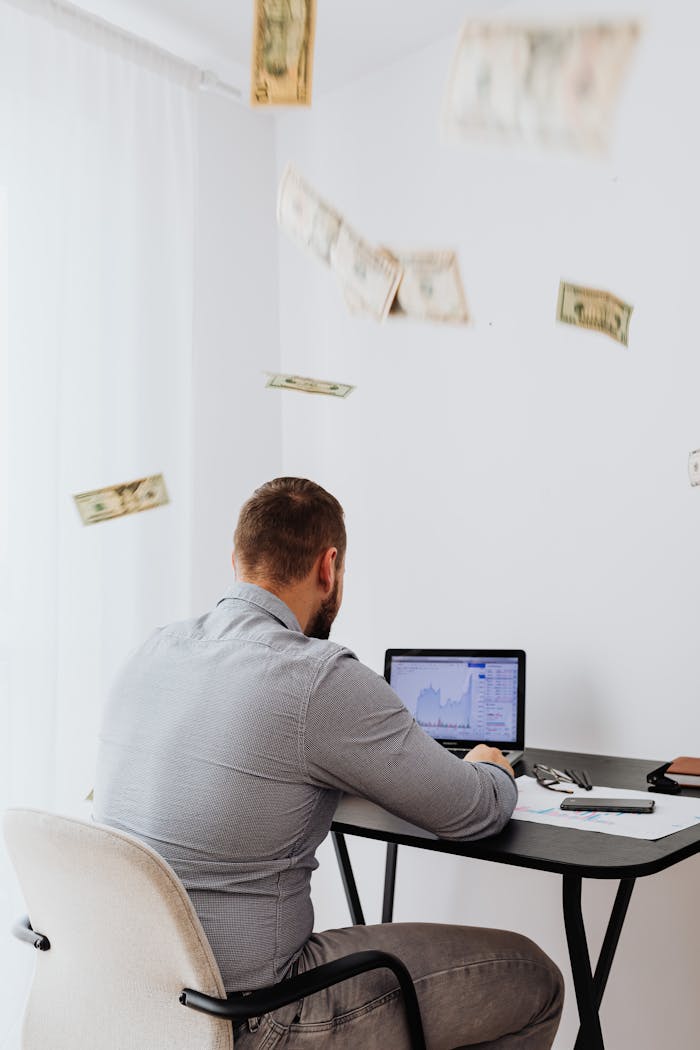 A man sits at a desk with a laptop and floating currency, symbolizing financial growth.
